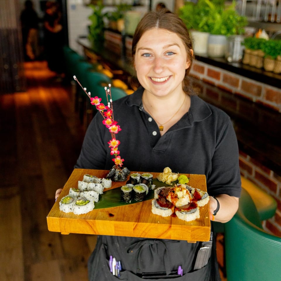 A smiling Miso server holds a wooden tray with various sushi rolls, ginger, and wasabi.